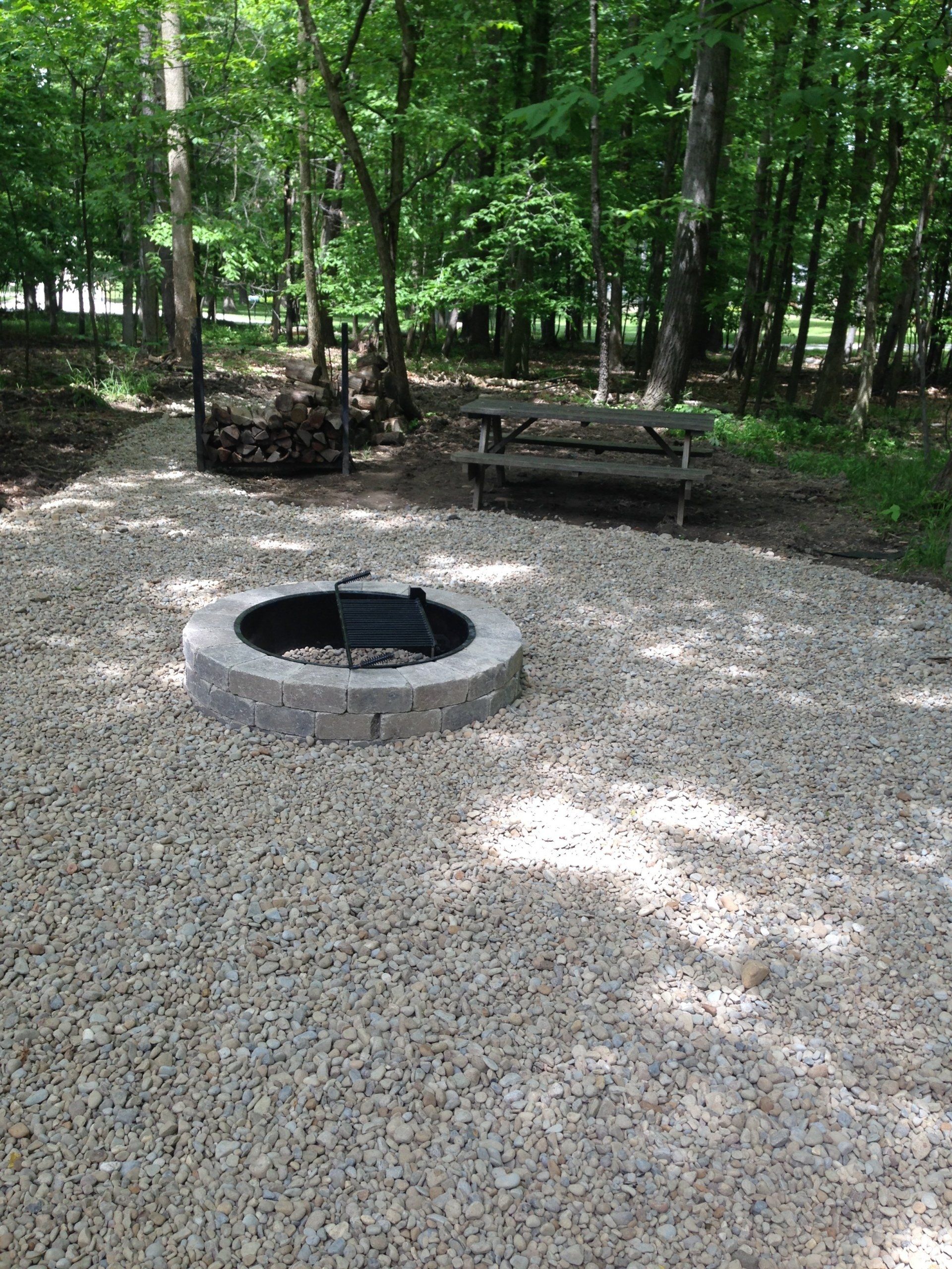 Circular fire pit on a gravel campsite in a wooded park with picnic tables in the background