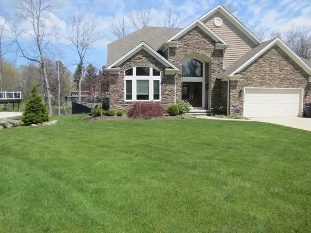 Suburban house with stone facade, white garage, and a large green lawn under a blue sky