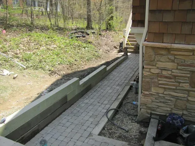 Outdoor stone walkway beside a house, with steps and a retaining wall along a wooded slope.