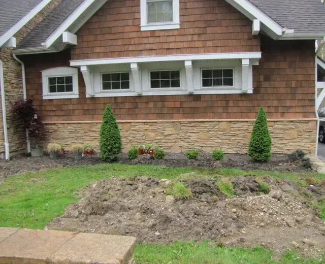 Front yard with fresh dirt and two small evergreen shrubs in front of a house with stone and wood siding