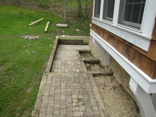 Brick walkway beside a house with a gravel strip and green lawn; construction materials in the yard