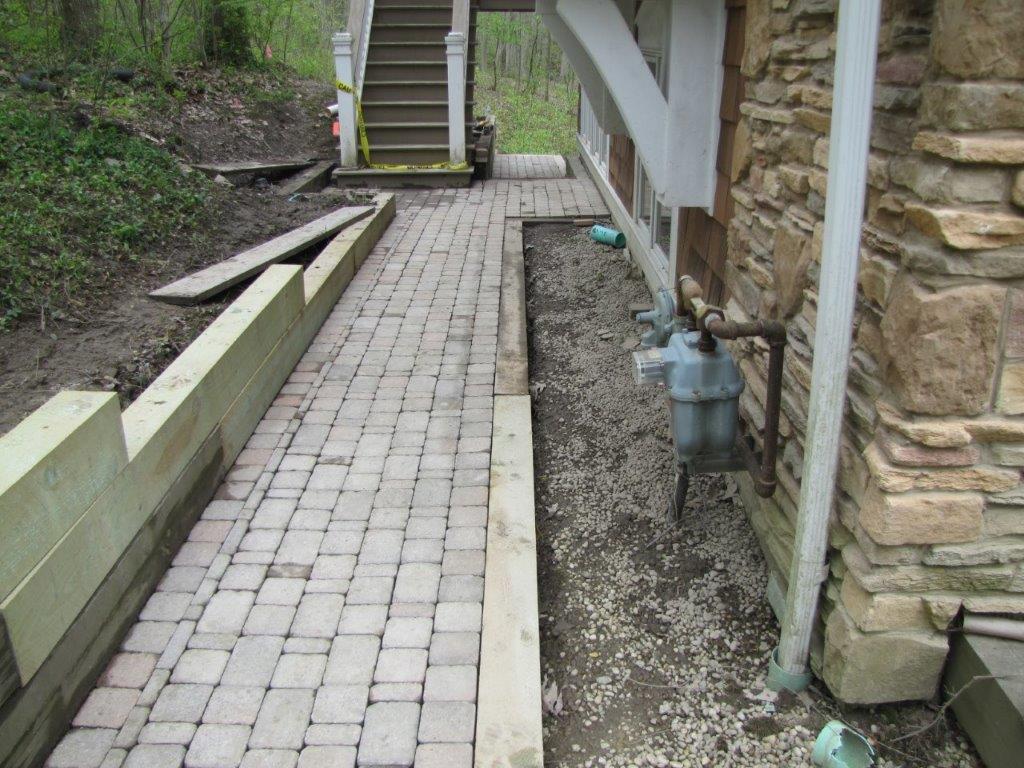 Narrow brick walkway beside a house and stone wall, with a steep path and steps ahead.