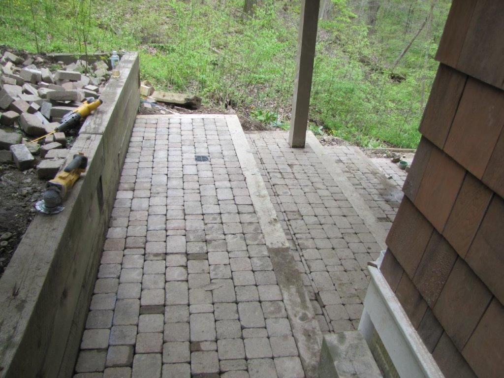 Brick patio walkway beside a shingled house, with a concrete wall and grassy slope.