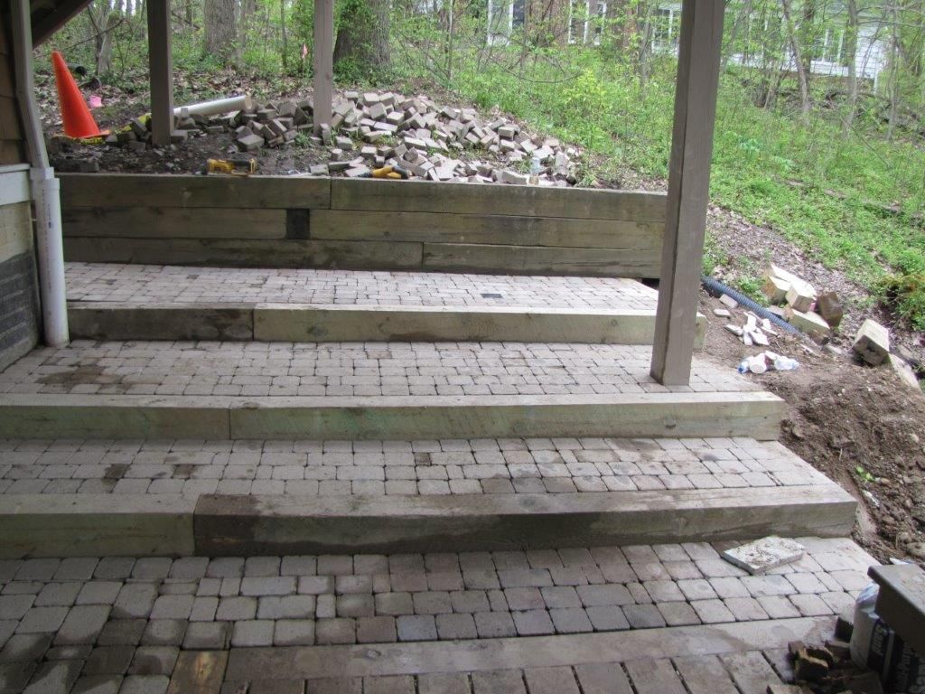Wooden porch with brick floor and steps, overlooking a wooded yard and rock pile.