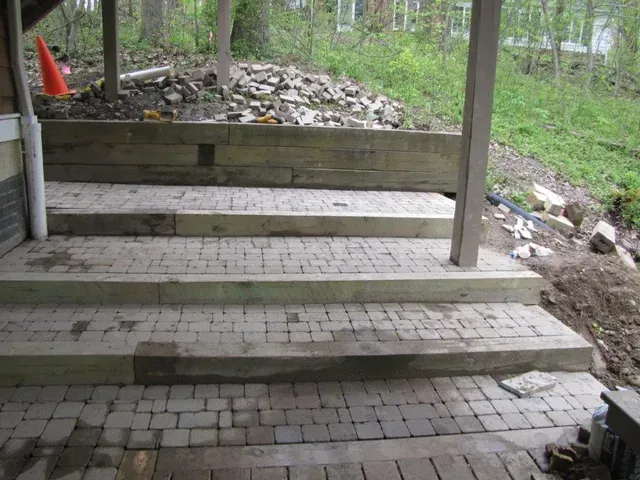Uneven brick patio steps under a covered outdoor area, with a stone pile and trees in the background.