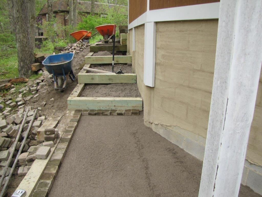 Exterior side yard with concrete steps, a wheelbarrow, and stacked building materials beside a house.