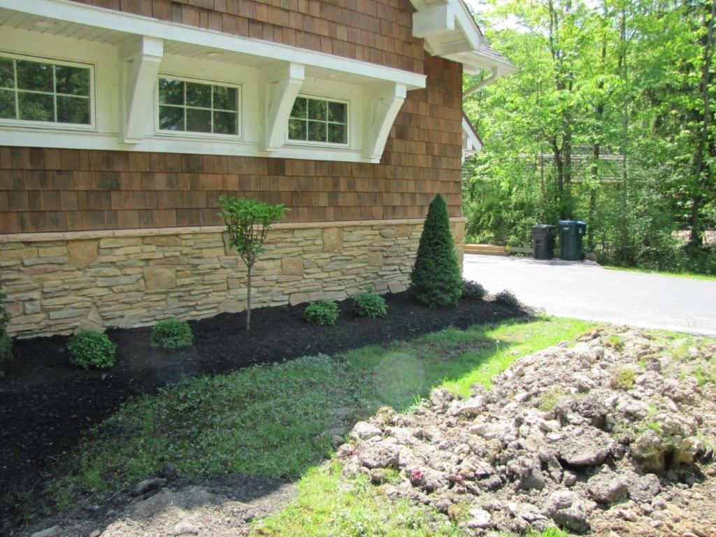 House exterior with stone foundation, white windows, garden bed, and a mulch pile beside a driveway