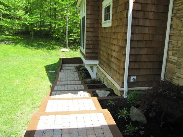 Wooden house side entrance with a narrow stone path and white accessibility ramp over a grassy yard.