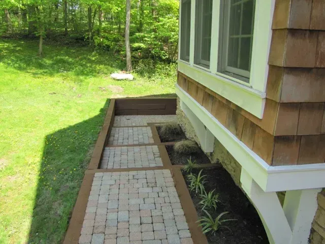 Brick walkway beside a house leading to a grassy yard and garden beds