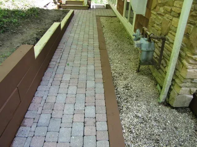 Brick walkway beside a house, bordered by gravel and a low brown retaining wall.