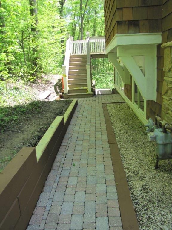 Narrow brick path beside a house leads to stairs into a green wooded area