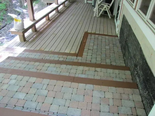 Patio walkway with wooden planks and brick pavers alongside a railing and building wall