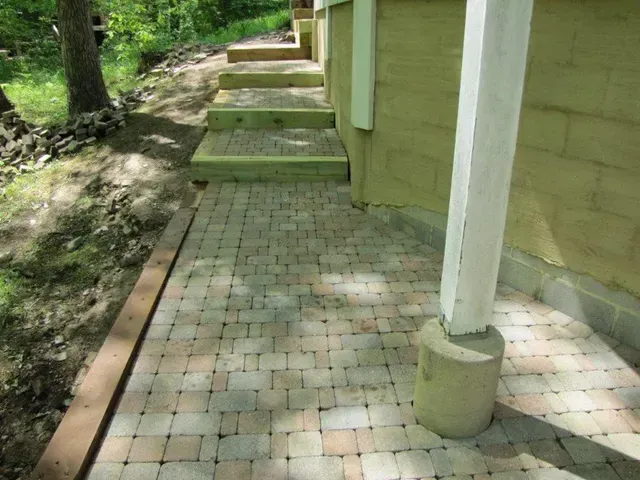 Brick patio walkway beside yellow house with white post and trees along the edge