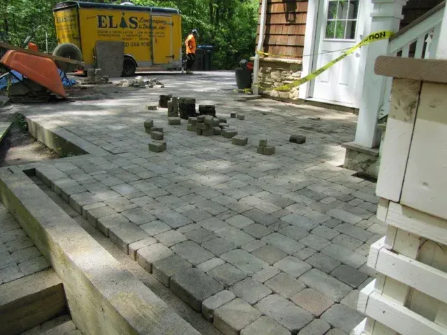 Stone patio under construction with scattered pavers and a yellow utility truck in the background