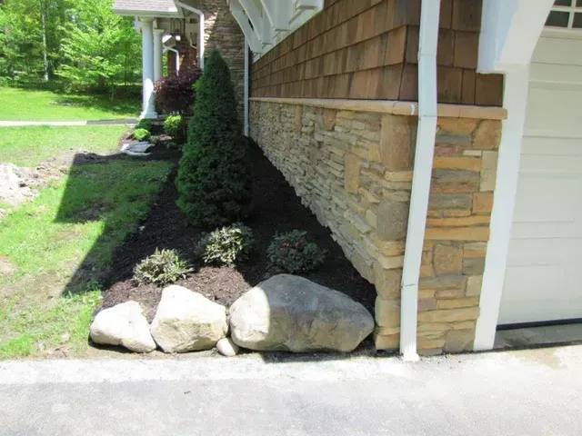 Landscaped house corner with shrubs, black mulch, and large rocks beside a driveway