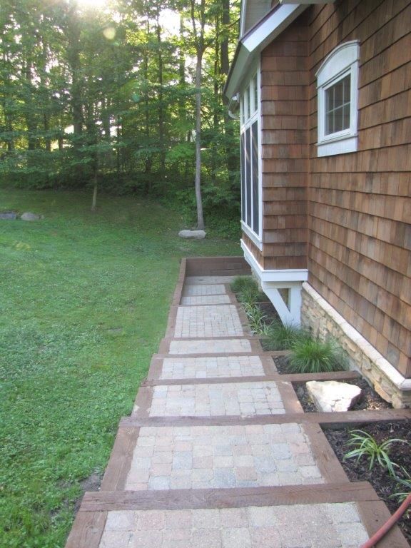 Side yard walkway with square pavers beside a brown shingled house and grassy lawn.
