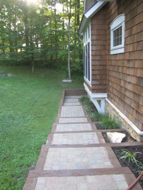 Brick walkway beside a shingled house leading to a grassy yard with trees