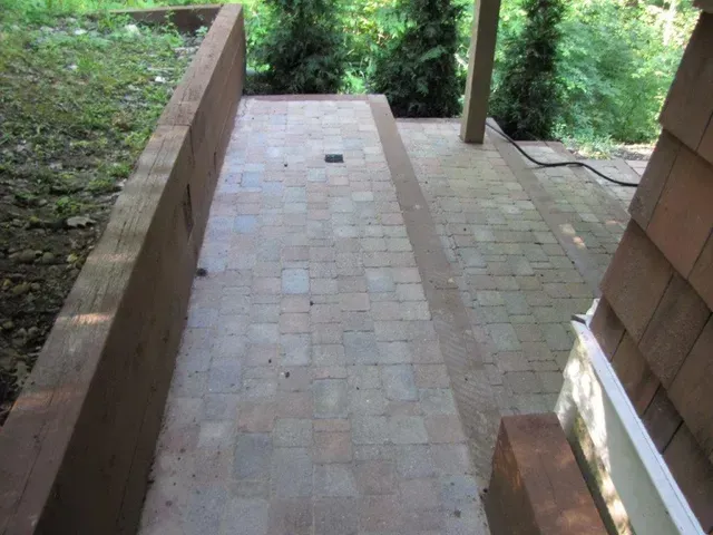 Brick-paved outdoor walkway with a narrow ramp beside a wooden railing and greenery