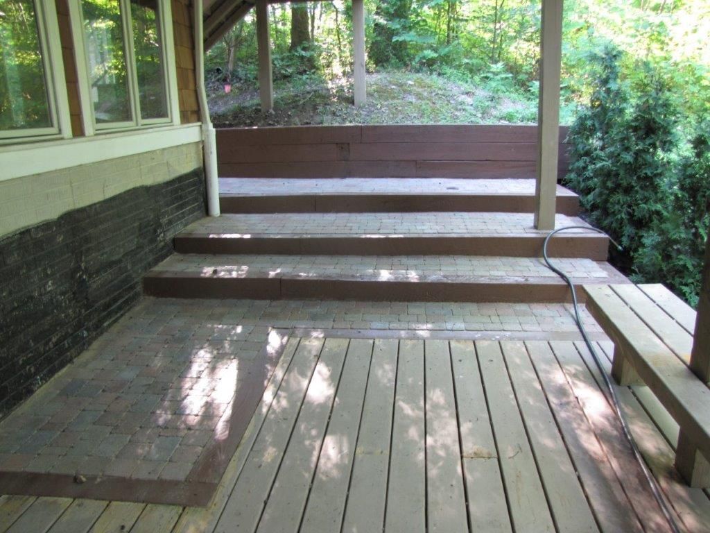 Wooden porch deck with steps, railing, and wooded yard beyond, beside a house wall