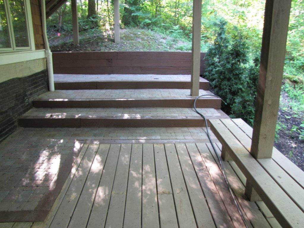 Wooden porch with steps, railing, and a bench-like side seat overlooking a wooded yard.