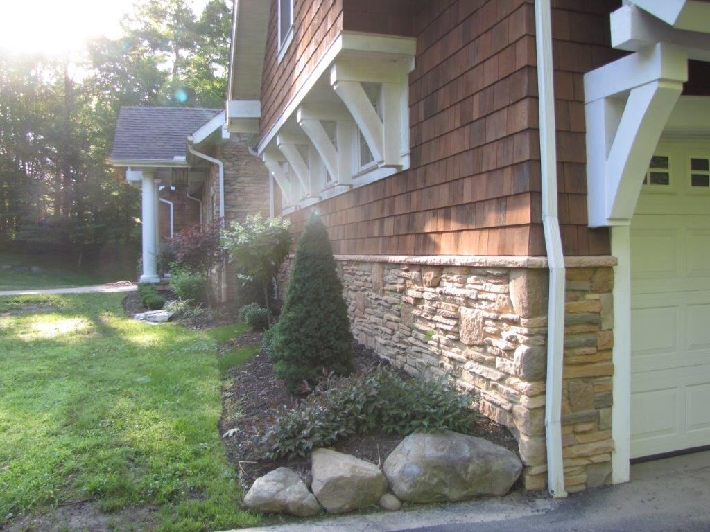 Side of a house with stone foundation, green lawn, shrubs, and a garage door in sunlight.