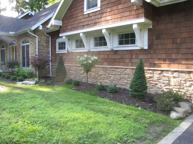 Suburban house exterior with shingled siding, stone facade, lawn, shrubs, and a driveway edge