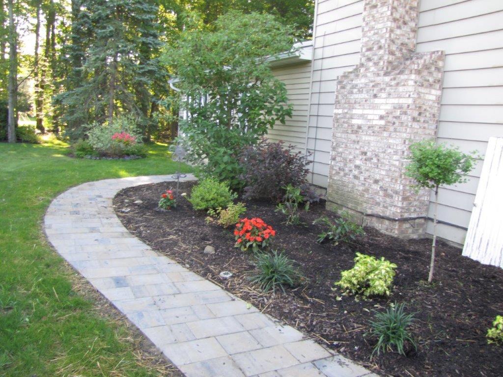 Curved stone walkway beside a house, bordered by a mulched flower bed with shrubs and red flowers.