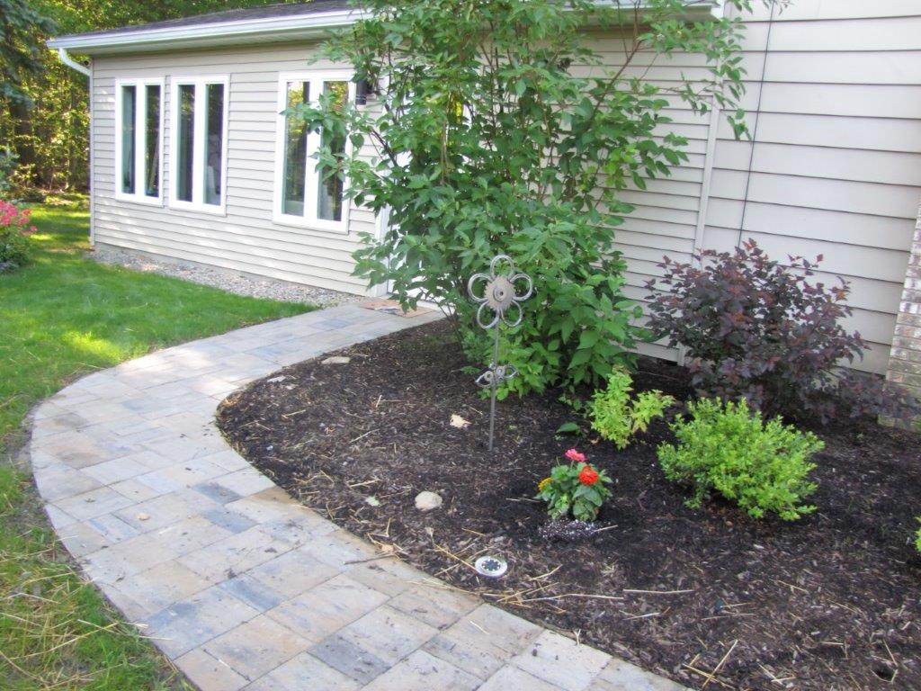Curved stone walkway beside a house with landscaped flower bed, shrubs, and a small tree