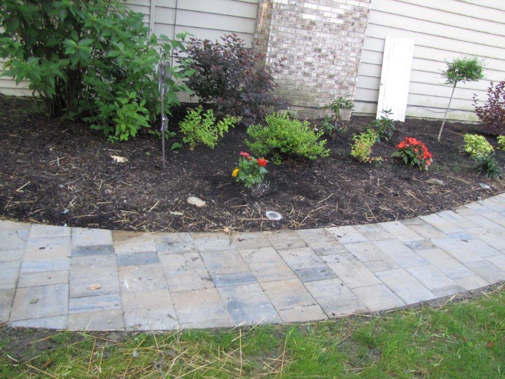 Curved stone walkway beside a mulched garden bed with shrubs and red flowers near a house exterior