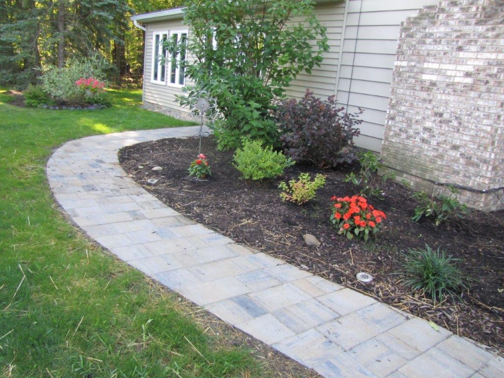 Curved stone walkway beside a house, bordered by a mulched garden bed with shrubs and red flowers.