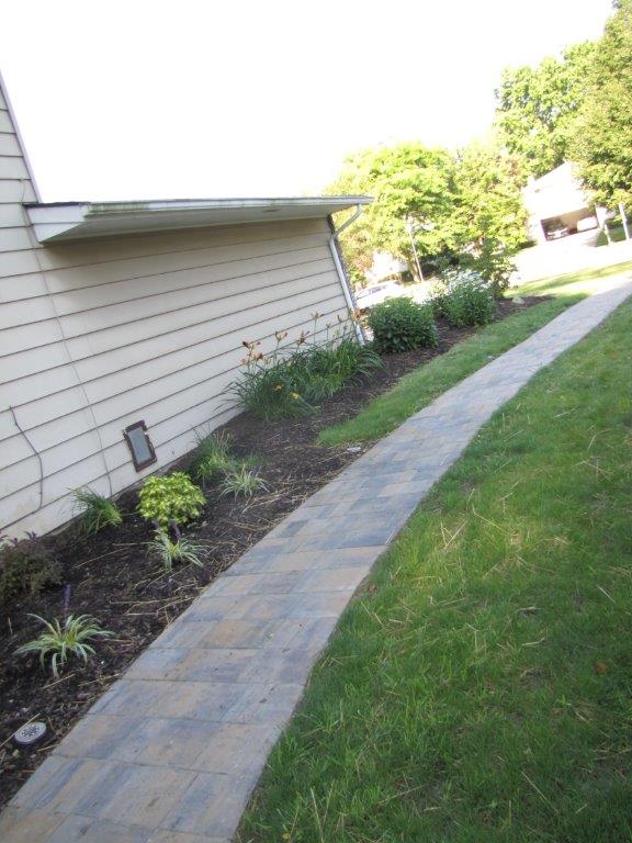 Side yard with a paved walkway beside a house, bordered by grass and low plants.