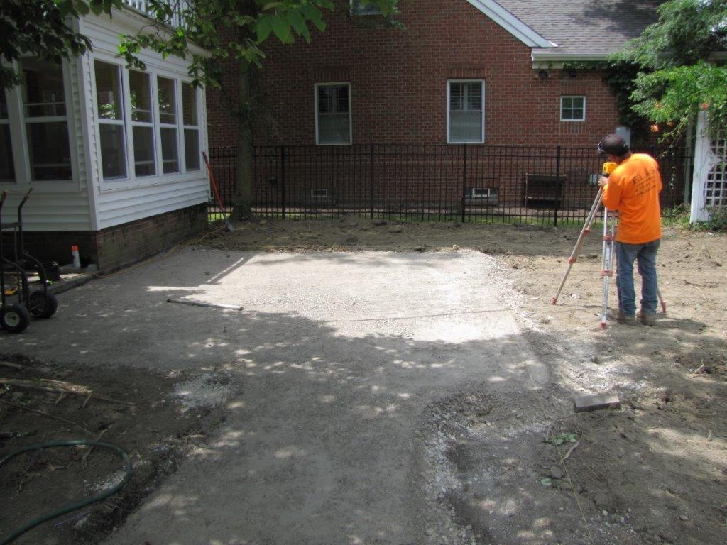 Worker in orange shirt leveling a gravel driveway beside a brick house and white porch.