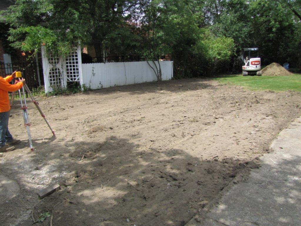 Dirt lot with trees and a white fence; worker in orange uses surveying equipment at left, SUV parked in background.