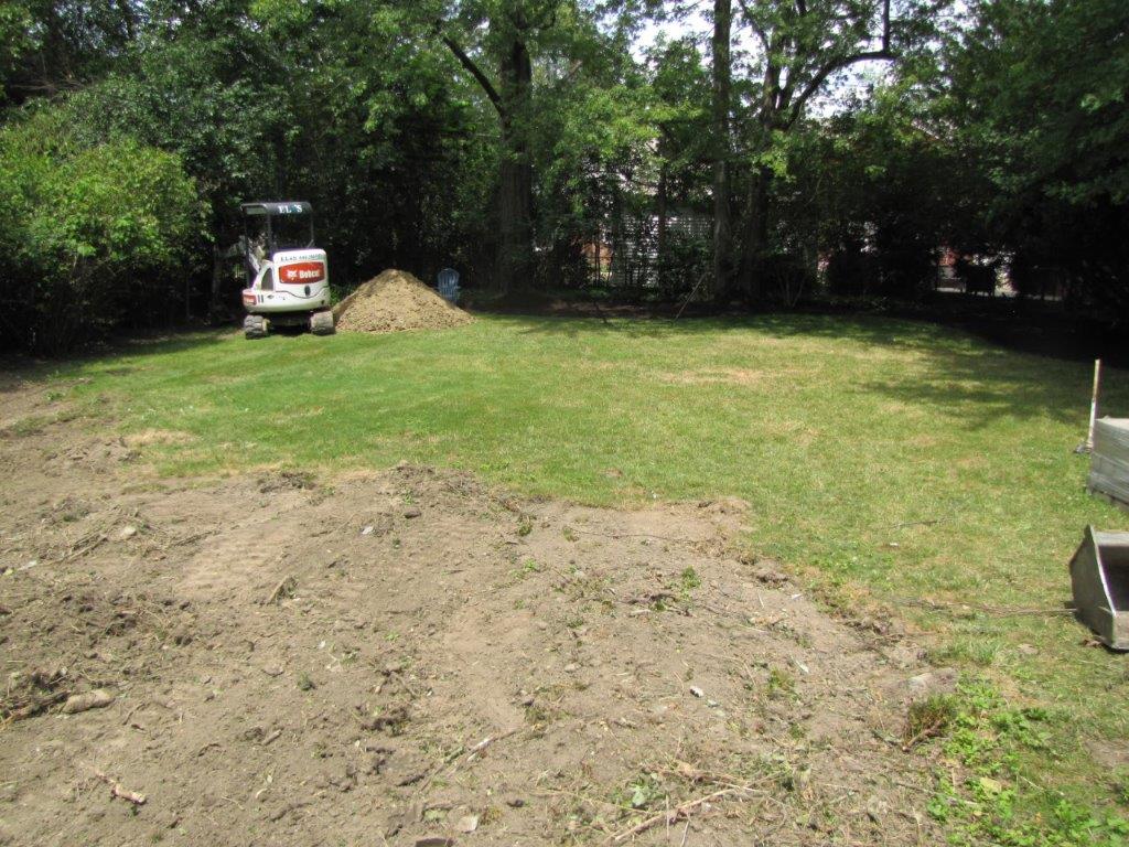 Dirt lot with a small skid steer, grass, trees, and a pile of soil in the background.