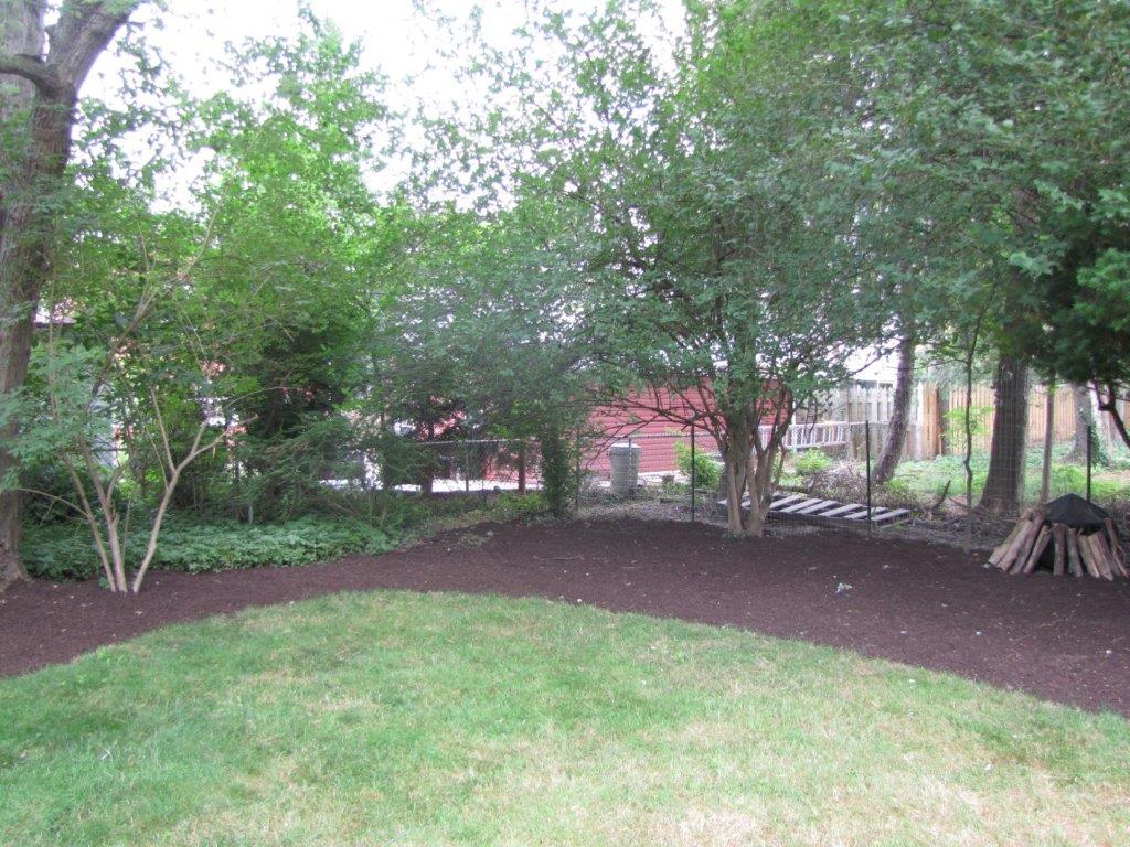 Grassy garden bordered by dark mulch and trees, with a red building in the background.