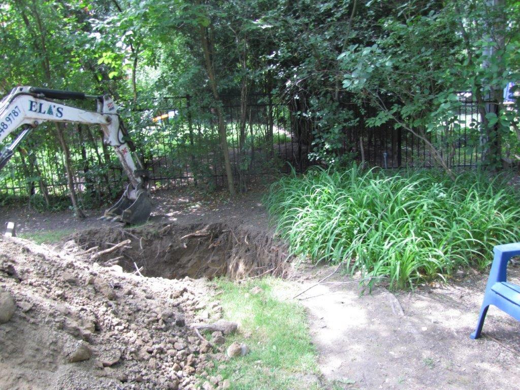 Backhoe excavating a dirt trench beside a wooded yard, with grass and a blue chair nearby
