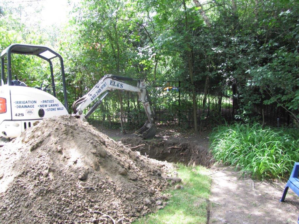 Excavator beside a dirt mound in a leafy yard or garden, with a path and fence in the background