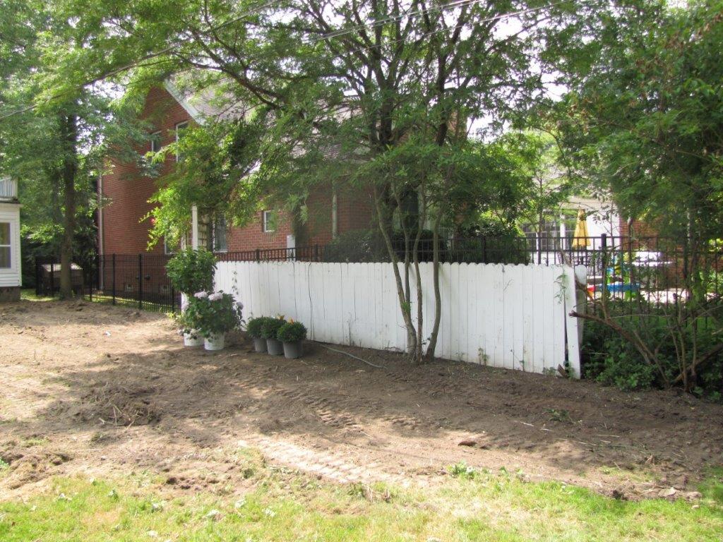 Tree-lined yard with a white fence beside a brick house on a dirt lot