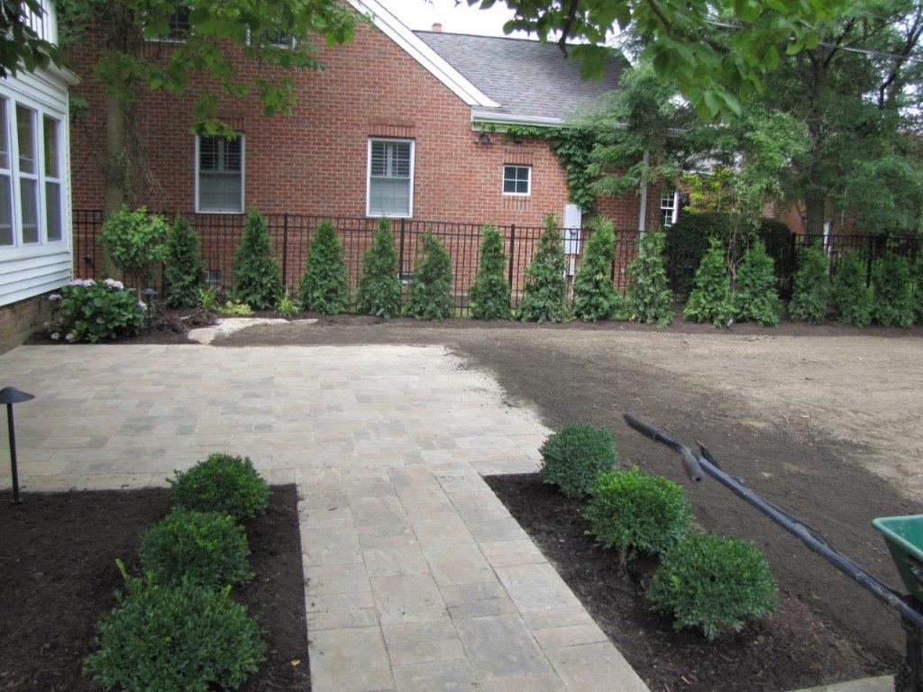 Brick house patio with stone walkway, small shrubs, and a gravel yard under trees