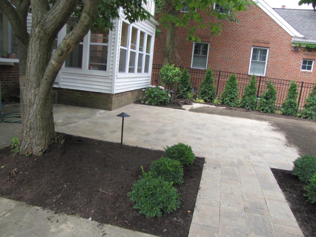 Front yard walkway with pavers, mulch beds, shrubs, and a tree beside a house and brick garage