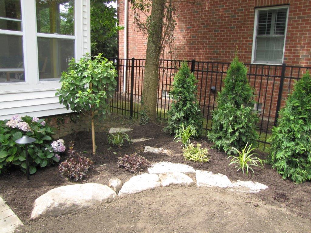 Front yard garden bed with shrubs, flowers, and white rocks beside a brick house and window