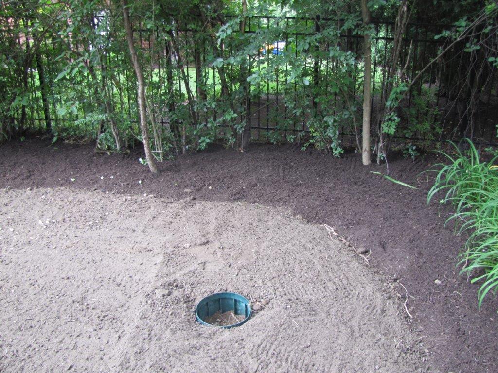 Circular ground opening in gravel near a wooded area, with a small blue container visible inside.