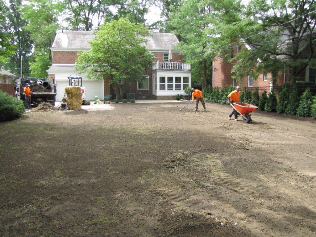 Workers spreading dirt in a yard beside a brick house and trees