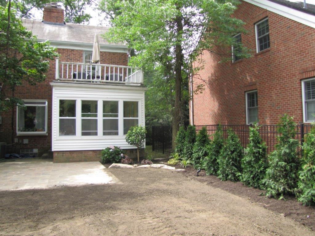 Backyard view of a brick house with a white porch and a dirt driveway beside shrubs