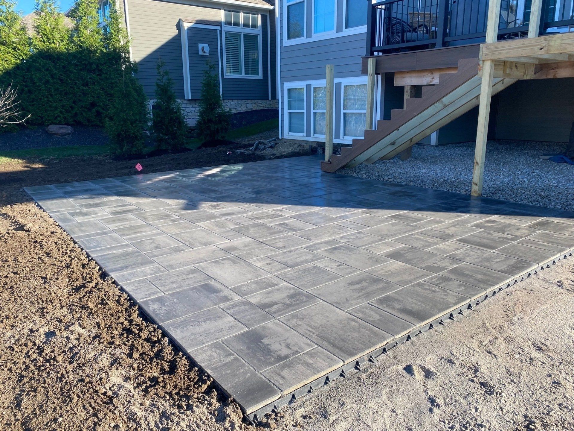 New paver patio beside a house, bordered by dirt and a wooden deck stairs.