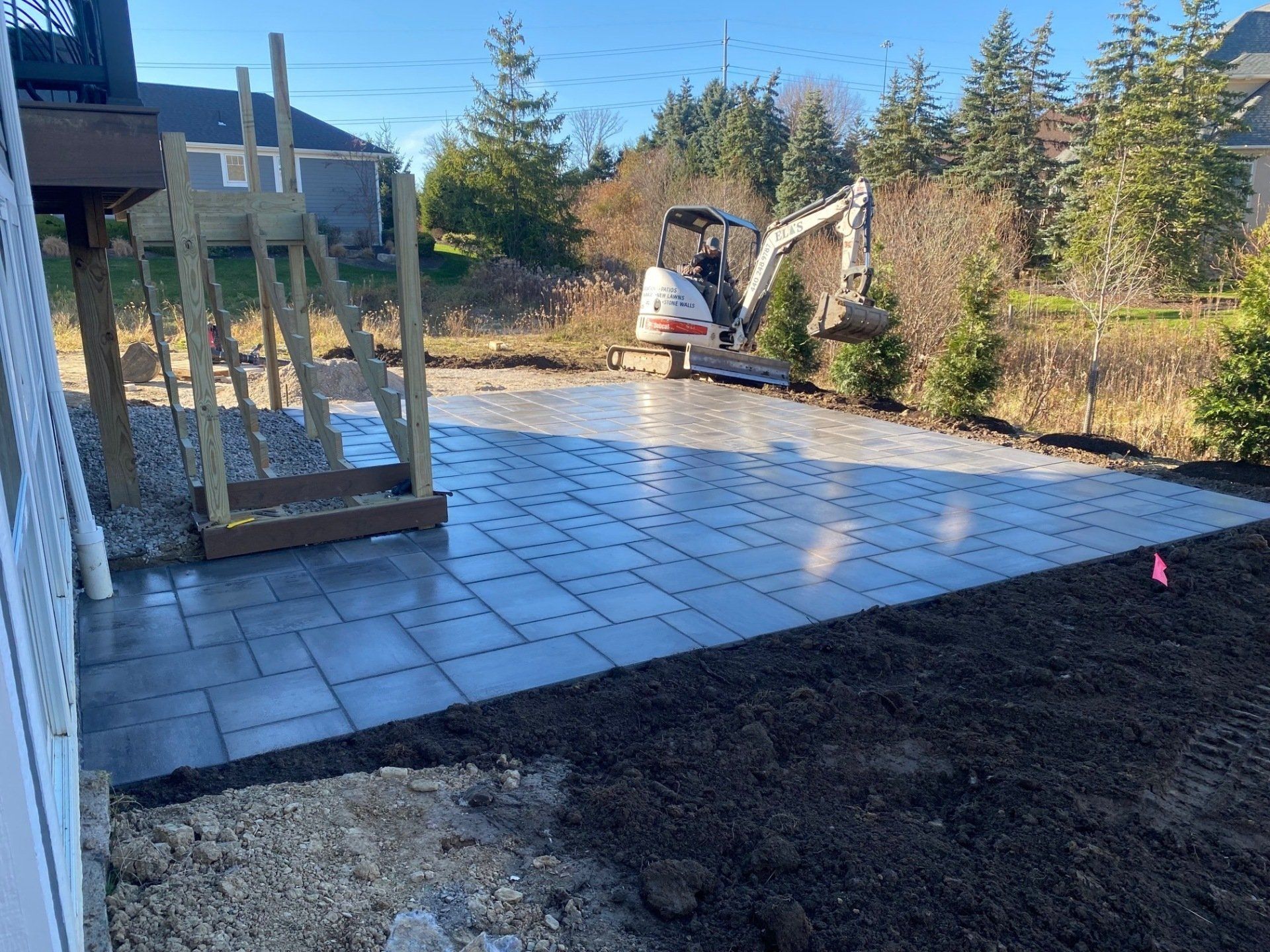 Partially paved backyard patio with a small excavator and unfinished gravel area beside a house