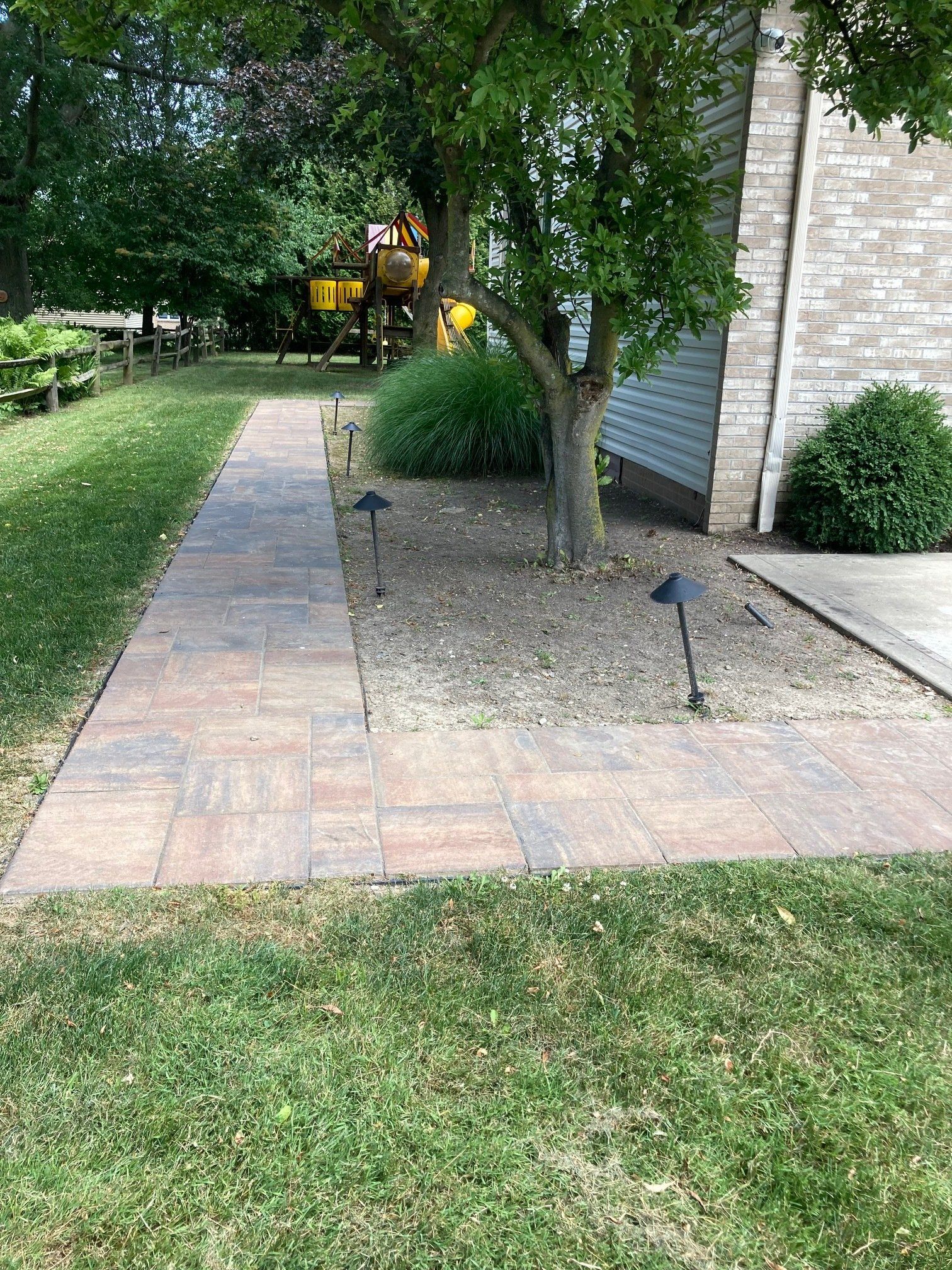 Brick walkway beside a house, lined with grass and trees, leading toward a yellow excavator in the distance