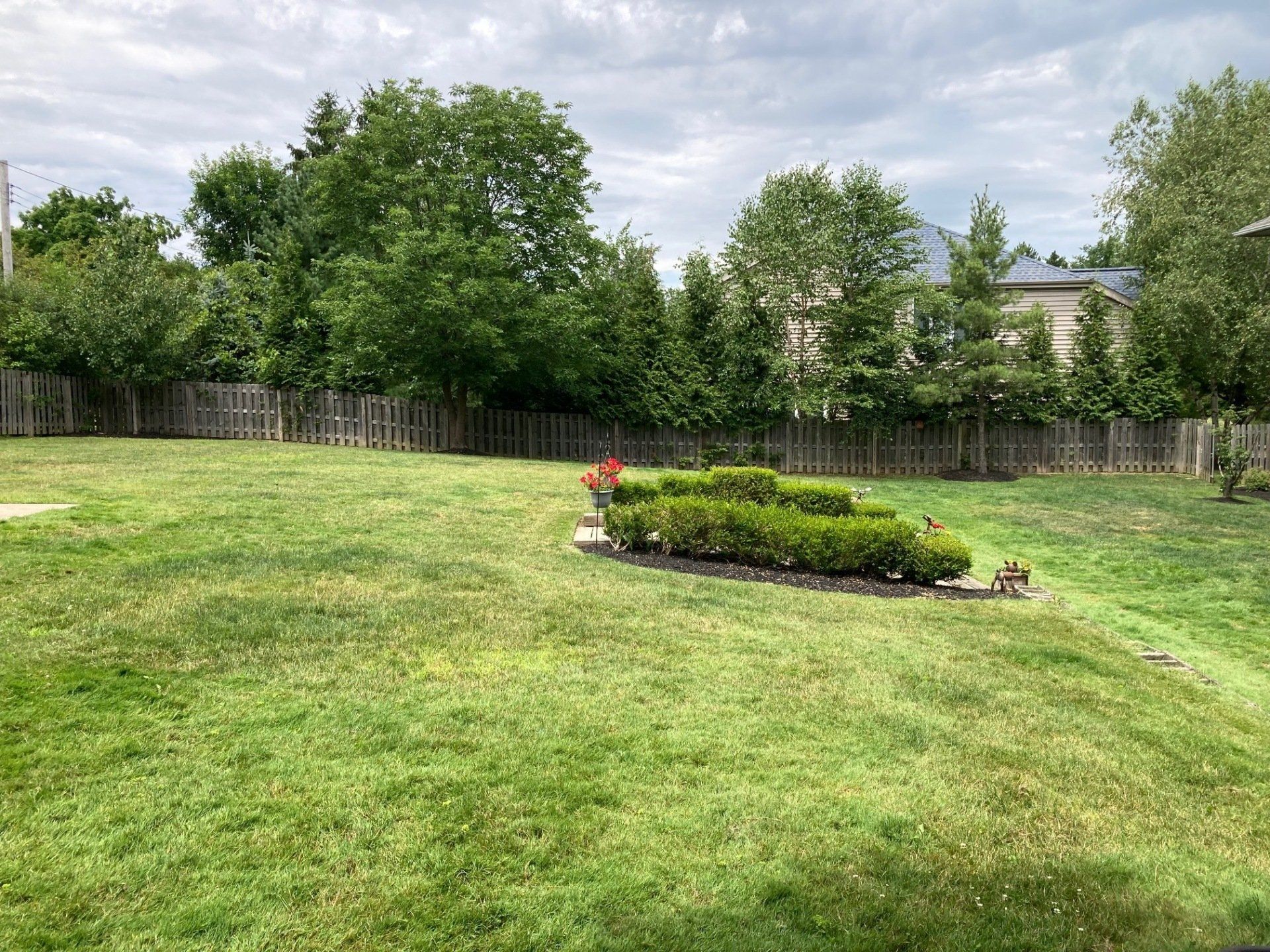 Grassy backyard with a small landscaped flower bed, trees, and a wooden fence under a cloudy sky
