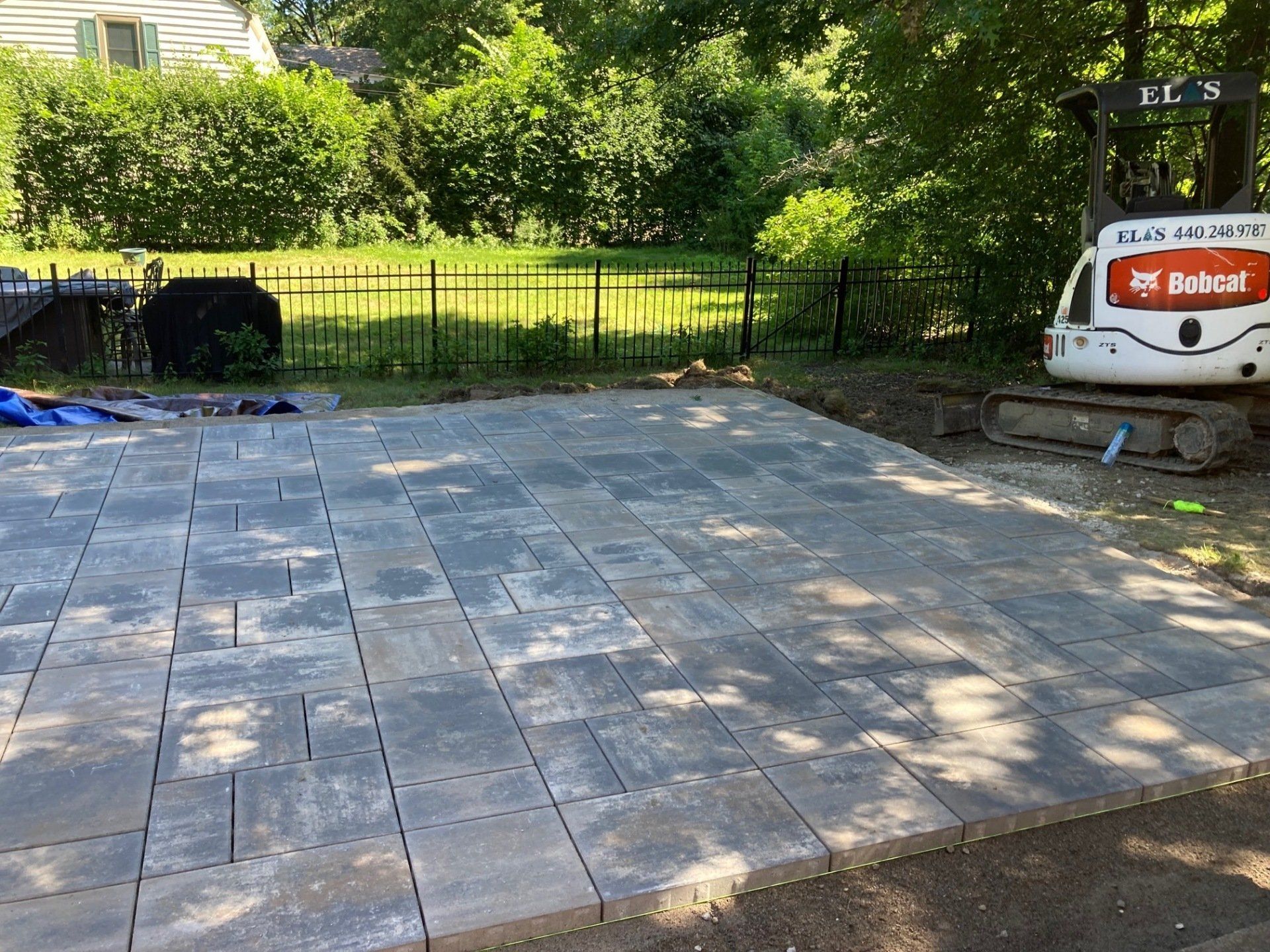 Patio with gray pavers in a backyard, bordered by grass and trees, with a Bobcat machine at right.