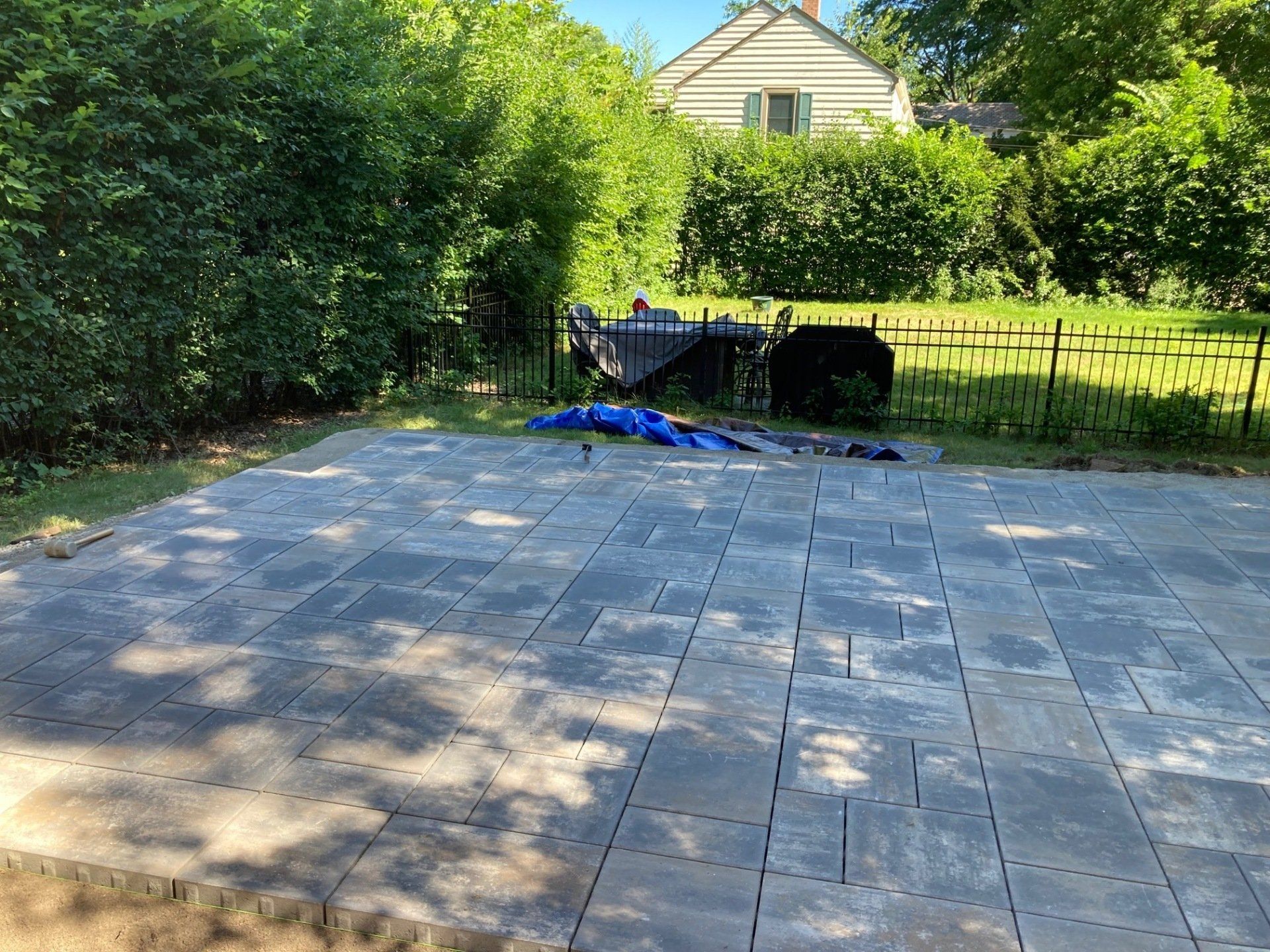 Stone patio with a wheelbarrow and blue tarp beside a hedge and black fence in a sunny backyard